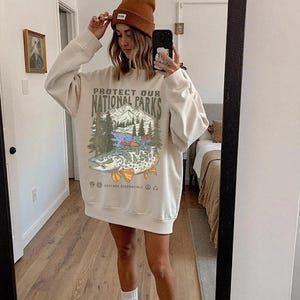 Puede incluir: Sudadera color crema con el texto "PROTECT OUR NATIONAL PARKS" y un gr&aacute;fico de un pez, monta&ntilde;as y &aacute;rboles. La persona lleva un gorro marr&oacute;n y calcetines blancos.