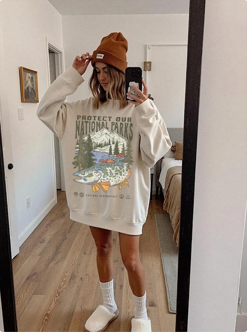Puede incluir: Sudadera color crema con el texto "PROTECT OUR NATIONAL PARKS" y un gr&aacute;fico de un pez, monta&ntilde;as y &aacute;rboles. La persona lleva un gorro marr&oacute;n y calcetines blancos.