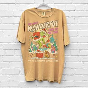 Puede incluir: Camiseta de color mostaza con un dibujo animado del Grinch vestido de Pap&aacute; Noel sobre una chimenea. El texto dice "THE MOST WONDERFUL TIME OF THE YEAR" y "WE'RE STEALING CHRISTMAS". El dise&ntilde;o incluye un perro y un pueblo festivo.