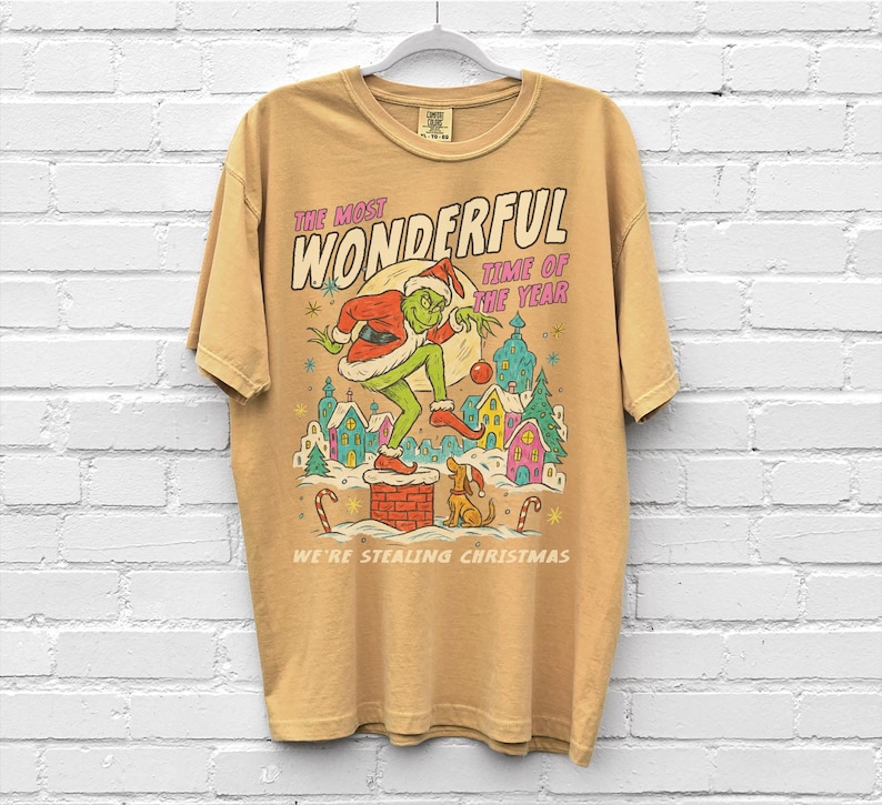 Puede incluir: Camiseta de color mostaza con un dibujo animado del Grinch vestido de Pap&aacute; Noel sobre una chimenea. El texto dice "THE MOST WONDERFUL TIME OF THE YEAR" y "WE'RE STEALING CHRISTMAS". El dise&ntilde;o incluye un perro y un pueblo festivo.