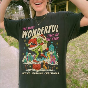 Puede incluir: Camiseta negra con un dise&ntilde;o del Grinch con gorro de Pap&aacute; Noel, robando la Navidad. El texto dice "The Most Wonderful Time of the Year" y "We're Stealing Christmas". El dise&ntilde;o incluye una colorida ilustraci&oacute;n de un pueblo nevado y bastones de caramelo.