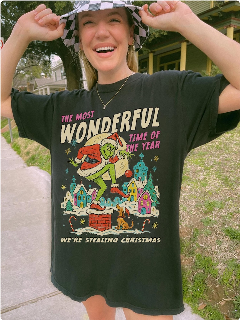 Puede incluir: Camiseta negra con un dise&ntilde;o del Grinch con gorro de Pap&aacute; Noel, robando la Navidad. El texto dice "The Most Wonderful Time of the Year" y "We're Stealing Christmas". El dise&ntilde;o incluye una colorida ilustraci&oacute;n de un pueblo nevado y bastones de caramelo.
