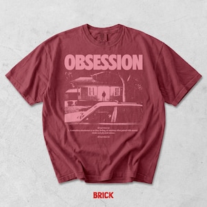 May include: A dark red short-sleeve t-shirt featuring a graphic print in light pink. The design includes the word OBSESSION in large capital letters above a house illustration with a car in the foreground and a dictionary definition of obsession.