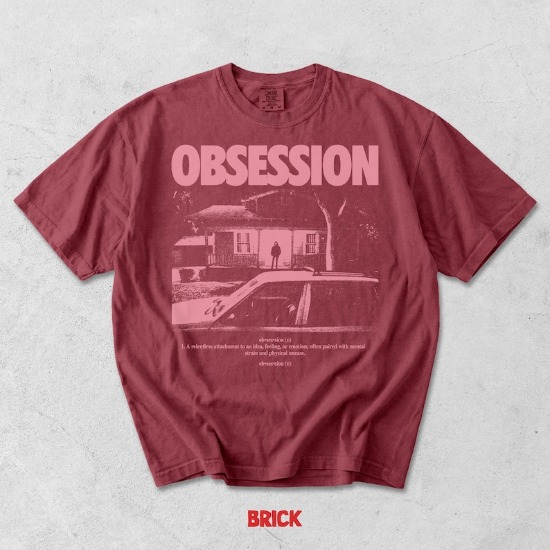 May include: A dark red short-sleeve t-shirt featuring a graphic print in light pink. The design includes the word OBSESSION in large capital letters above a house illustration with a car in the foreground and a dictionary definition of obsession.