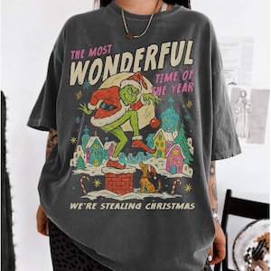 Puede incluir: Camiseta gris oscuro con un gr&aacute;fico tem&aacute;tico navide&ntilde;o. El dise&ntilde;o presenta un personaje verde con un gorro de Pap&aacute; Noel, un perro y casas coloridas. El texto en la camiseta dice "THE MOST WONDERFUL TIME OF THE YEAR" y "WE'RE STEALING CHRISTMAS."