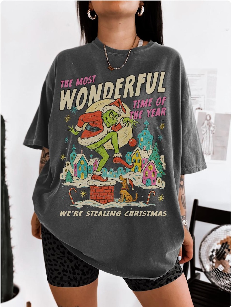 Puede incluir: Camiseta gris oscuro con un gr&aacute;fico tem&aacute;tico navide&ntilde;o. El dise&ntilde;o presenta un personaje verde con un gorro de Pap&aacute; Noel, un perro y casas coloridas. El texto en la camiseta dice "THE MOST WONDERFUL TIME OF THE YEAR" y "WE'RE STEALING CHRISTMAS."