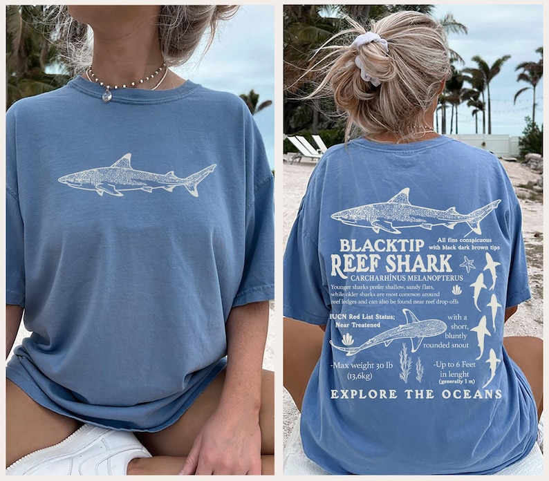May include: A light blue t-shirt with a white shark graphic on the front. The back of the shirt has a detailed illustration of a Blacktip Reef Shark, scientific details, and the words "Explore the Oceans".