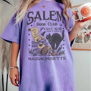 May include: A violet-colored t-shirt with the text "SALEM BOOK CLUB" and a graphic of a black cat, books, and a scroll. The shirt also includes the text "EST. 1626" and "MASSACHUSETTS".