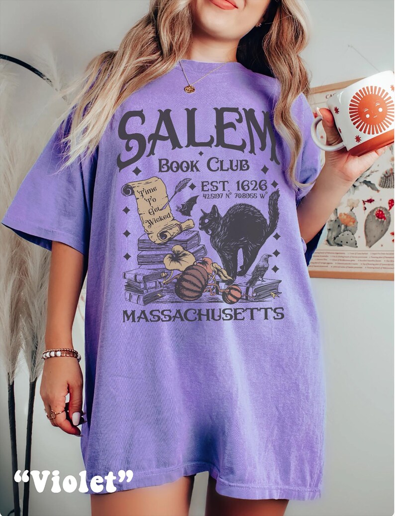 May include: A violet-colored t-shirt with the text "SALEM BOOK CLUB" and a graphic of a black cat, books, and a scroll. The shirt also includes the text "EST. 1626" and "MASSACHUSETTS".