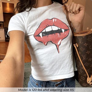 Puede incluir: Una camiseta blanca de manga corta con un gráfico rojo desgastado de labios con colmillos blancos y un efecto de goteo. La camiseta se combina con vaqueros azules y un bolso de hombro marrón con estampado. El modelo pesa 54 kg y usa la talla XS.