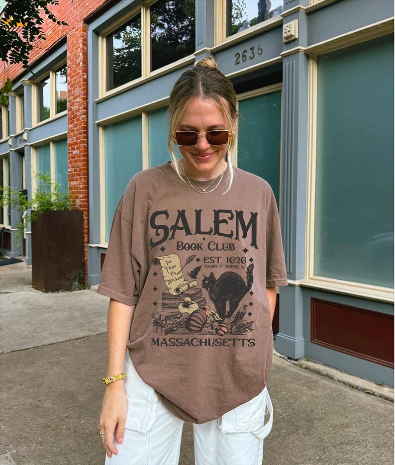 May include: A brown t-shirt with the text "Salem Book Club" and a graphic of a black cat, books, and a pumpkin. The shirt also includes the text "Est. 1626" and "Massachusetts". The shirt is worn with white pants.