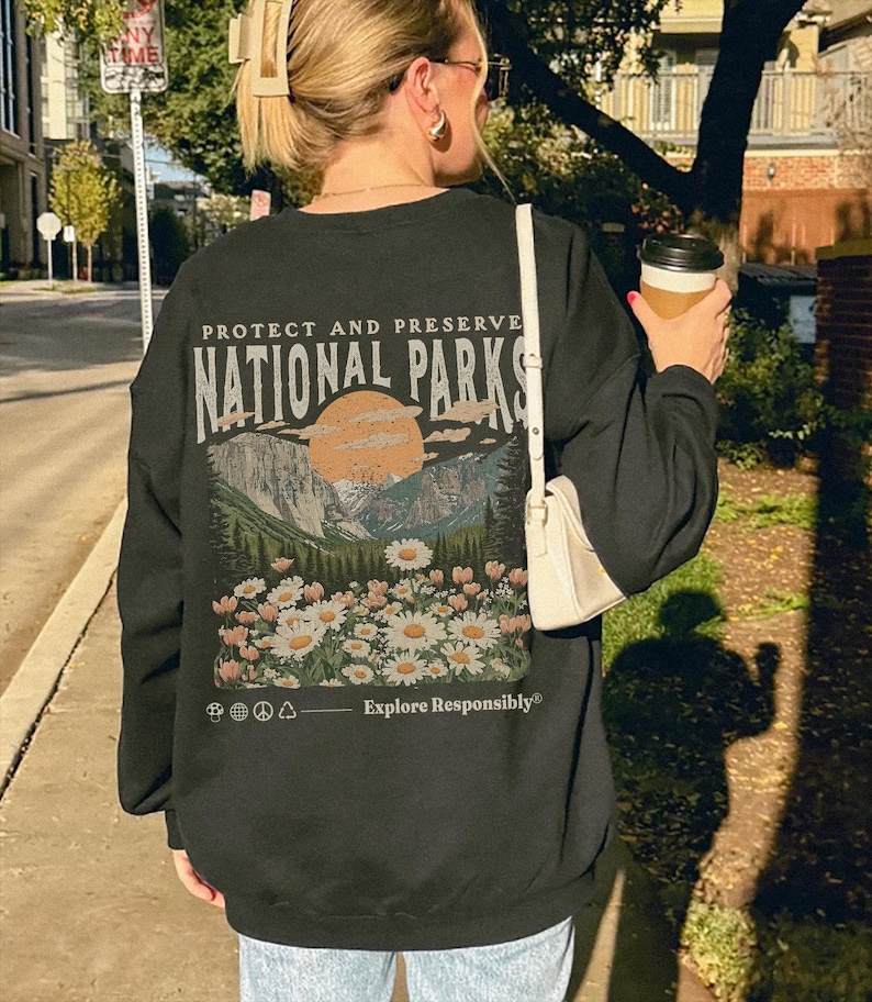Op de afbeelding: Een zwart sweatshirt met ronde hals en een rugprint met de tekst "PROTECT AND PRESERVE NATIONAL PARKS" boven een illustratie van bergen, een zon en een veld met madeliefjes, met de tekst "Explore Responsibly" en kleine iconen eronder.