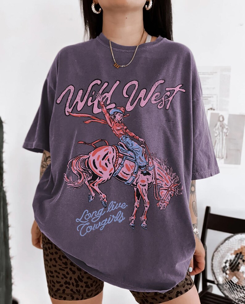 May include: A dark purple short-sleeve t-shirt featuring a graphic print of a person riding a bucking horse. The text Wild West is printed in pink above the graphic, and the text Long live cowgirls is printed in light blue below the horse.