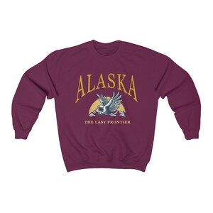 Peut inclure: Sweat-shirt bordeaux avec un graphique dor&eacute; d'un aigle volant au-dessus des montagnes avec le texte "Alaska The Last Frontier".
