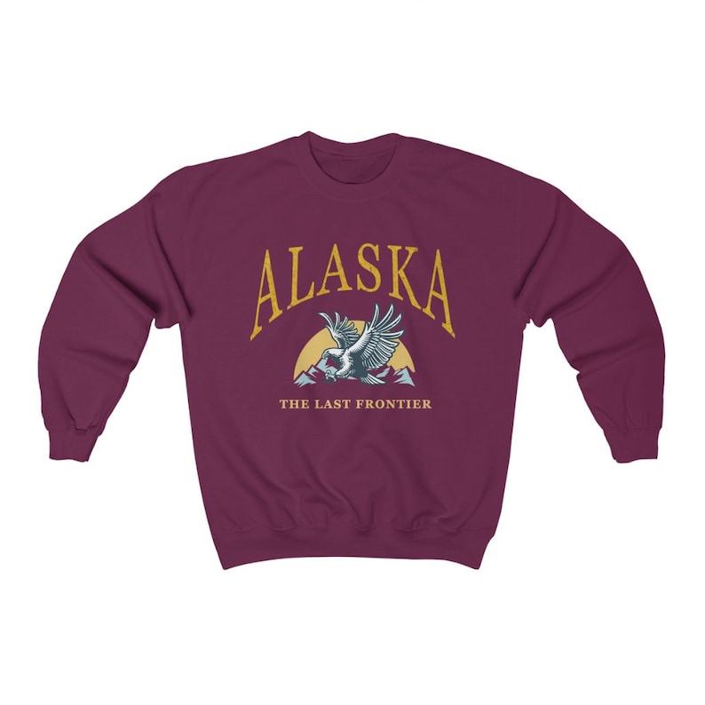 Peut inclure: Sweat-shirt bordeaux avec un graphique dor&eacute; d'un aigle volant au-dessus des montagnes avec le texte "Alaska The Last Frontier".