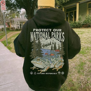 May include: A black hooded sweatshirt featuring a graphic on the back with the text "PROTECT OUR NATIONAL PARKS" above an illustration of mountains, pine trees, a river, and a large spotted frog, with "EXPLORE RESPONSIBLY" printed below.