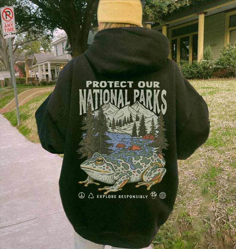 May include: A black hooded sweatshirt featuring a graphic on the back with the text "PROTECT OUR NATIONAL PARKS" above an illustration of mountains, pine trees, a river, and a large spotted frog, with "EXPLORE RESPONSIBLY" printed below.