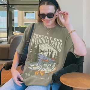 May include: A green t-shirt with a graphic print of a mountain scene and a fish. The text on the shirt reads "Protect Our National Parks" and "Explore Responsibly".