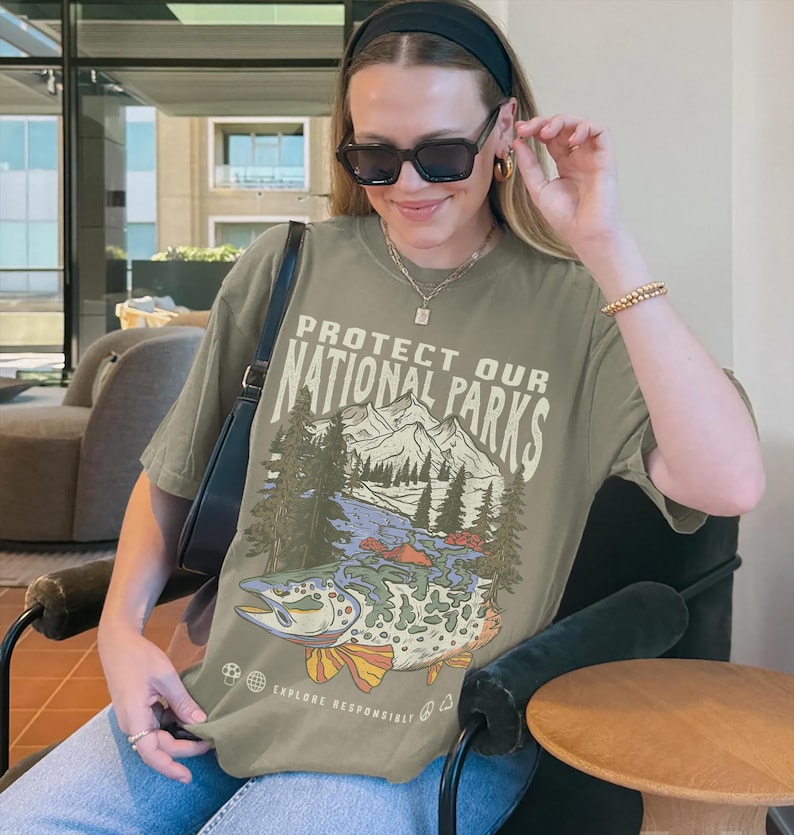May include: A green t-shirt with a graphic print of a mountain scene and a fish. The text on the shirt reads "Protect Our National Parks" and "Explore Responsibly".
