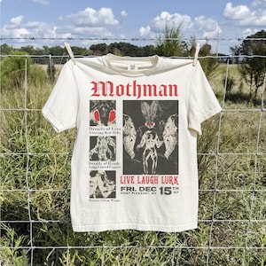 Könnte beinhalten: Ein cremefarbenes T-Shirt mit einer schwarz-roten Grafik des Mothman. Das Design enthält den Text Mothman, Live Laugh Lurk und Fri. Dec 15th '67 Point Pleasant, WV, sowie drei kleinere Felder, die leuchtend rote Augen, lange Klauenfinger und massive, stille Flügel zeigen.