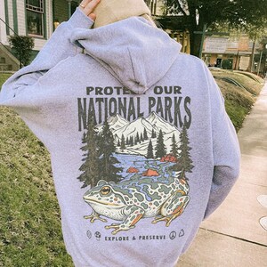May include: A light purple hooded sweatshirt featuring a graphic on the back with the text PROTECT OUR NATIONAL PARKS and EXPLORE & PRESERVE. The design includes a large frog, a mountain range, pine trees, a river, a mushroom, and a peace sign.