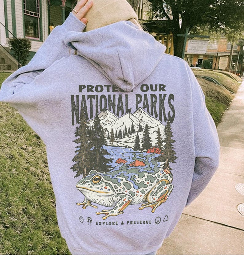 May include: A light purple hooded sweatshirt featuring a graphic on the back with the text PROTECT OUR NATIONAL PARKS and EXPLORE & PRESERVE. The design includes a large frog, a mountain range, pine trees, a river, a mushroom, and a peace sign.