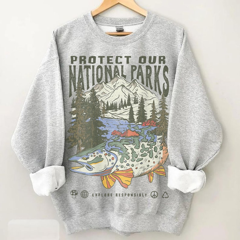 Puede incluir: Una sudadera gris con mangas dobladas que presenta un gr&aacute;fico de un pez con una escena de monta&ntilde;a y lago. El texto "PROTECT OUR NATIONAL PARKS" est&aacute; impreso encima de la imagen, con "EXPLORE RESPONSIBLY" debajo.