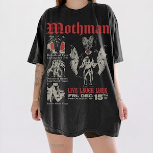 Può includere: Una t-shirt nero antracite con una stampa grafica che riporta il testo Mothman, Live Laugh Lurk e Fri. Dec 15th '67 Point Pleasant, WV. Il design include illustrazioni di occhi rossi luminosi, mani artigliate e ali massicce.