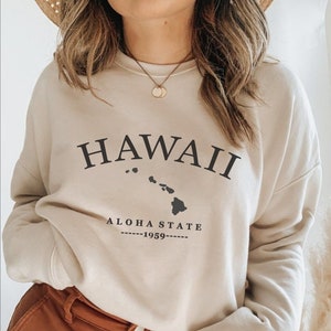Puede incluir: Una sudadera beige con un estampado gráfico negro del estado de Hawái, el texto "HAWAII", "ALOHA STATE" y "1959".