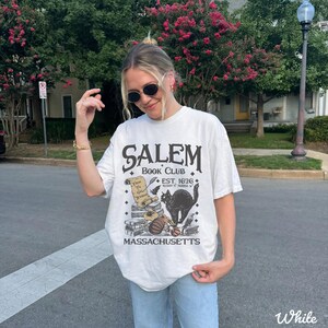 May include: White t-shirt with a graphic design featuring the text "Salem Book Club" and a black cat illustration. The design includes the text "Est. 1626" and "Massachusetts". The shirt is a casual, oversized fit.