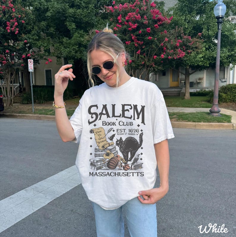 May include: White t-shirt with a graphic design featuring the text "Salem Book Club" and a black cat illustration. The design includes the text "Est. 1626" and "Massachusetts". The shirt is a casual, oversized fit.