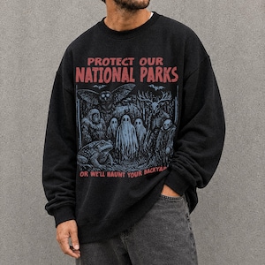 Puede incluir: Una sudadera negra de cuello redondo con un estampado gráfico que incluye el texto en rojo "PROTECT OUR NATIONAL PARKS" y "OR WE'LL HAUNT YOUR BACKYARD". La ilustración muestra un búho, una rana, fantasmas y un esqueleto en un bosque.