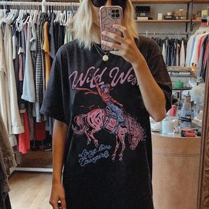 Puede incluir: Una camiseta negra de manga corta con un estampado gr&aacute;fico de una vaquera montando un caballo encabritado. El texto "Wild West" aparece en rosa sobre la ilustraci&oacute;n y la frase "Long live Cowgirls" est&aacute; impresa en azul debajo del caballo.