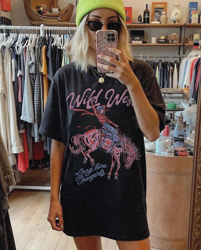 Puede incluir: Una camiseta negra de manga corta con un estampado gr&aacute;fico de una vaquera montando un caballo encabritado. El texto "Wild West" aparece en rosa sobre la ilustraci&oacute;n y la frase "Long live Cowgirls" est&aacute; impresa en azul debajo del caballo.