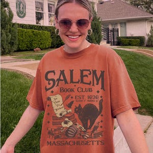 May include: Rust-colored t-shirt with the text "SALEM BOOK CLUB" and the words "EST. 1626" and "MASSACHUSETTS". The graphic includes a black cat, books, a scroll, and a pumpkin. The shirt is worn by a person wearing sunglasses.