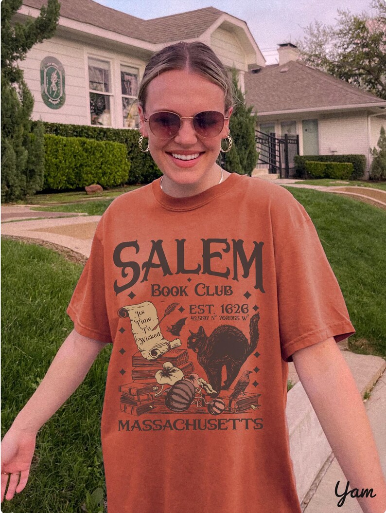 May include: Rust-colored t-shirt with the text "SALEM BOOK CLUB" and the words "EST. 1626" and "MASSACHUSETTS". The graphic includes a black cat, books, a scroll, and a pumpkin. The shirt is worn by a person wearing sunglasses.