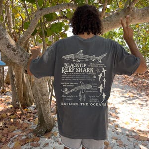 May include: Charcoal grey t-shirt with a graphic of a Blacktip Reef Shark, scientific details, and the words "EXPLORE THE OCEANS." The design includes shark illustrations and text describing the species' features.