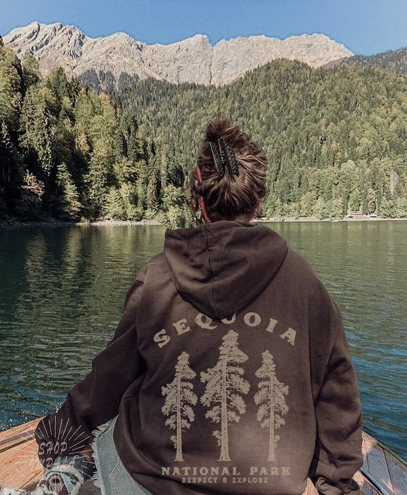 May include: A brown hoodie with a graphic of three sequoia trees and the text "Sequoia National Park Respect & Explore". The person wearing the hoodie is standing in front of a lake and mountains.