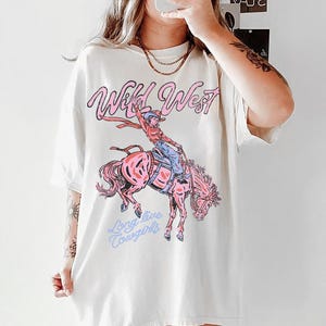 Puede incluir: Una camiseta de manga corta color blanco roto con un estampado gr&aacute;fico de una vaquera montando un caballo que corcovea. El texto "Wild West" aparece en rosa sobre la ilustraci&oacute;n y "Long live Cowgirls" est&aacute; impreso en azul claro debajo.