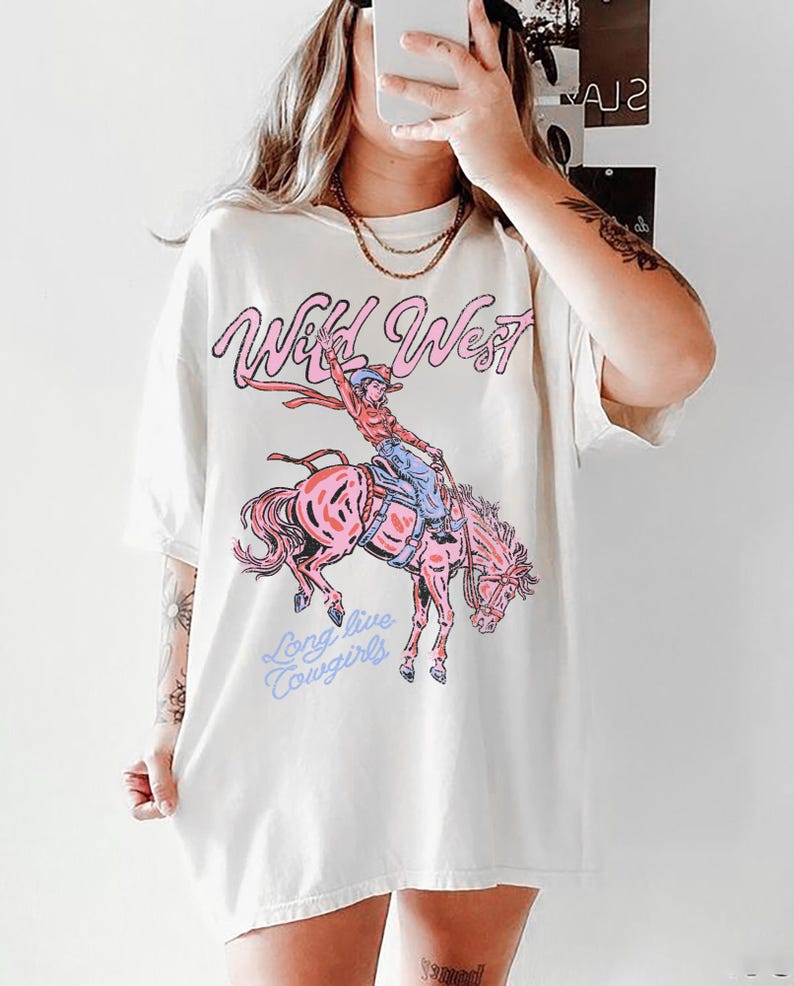 Puede incluir: Una camiseta de manga corta color blanco roto con un estampado gr&aacute;fico de una vaquera montando un caballo que corcovea. El texto "Wild West" aparece en rosa sobre la ilustraci&oacute;n y "Long live Cowgirls" est&aacute; impreso en azul claro debajo.