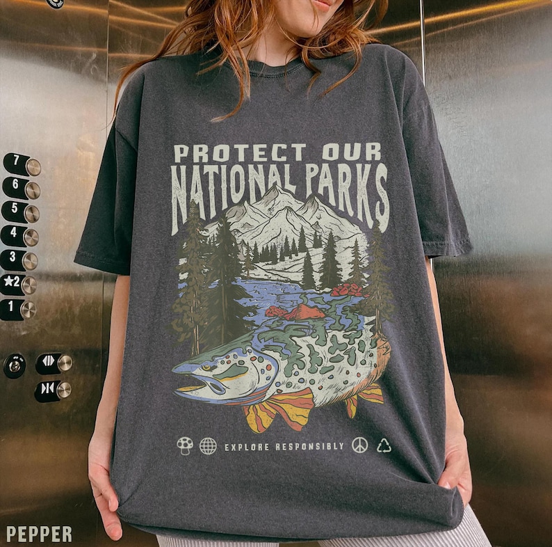 May include: A dark gray t-shirt with a graphic print of a fish swimming in a lake with mountains in the background. The text "PROTECT OUR NATIONAL PARKS" is printed above the image. The text "EXPLORE RESPONSIBLY" is printed below the image.