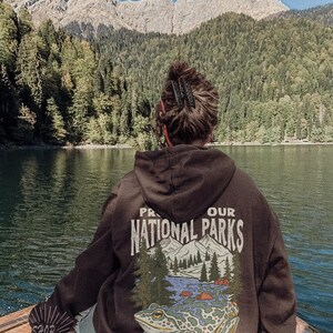 May include: A dark brown hooded sweatshirt featuring a graphic on the back with the text PROTECT OUR NATIONAL PARKS and EXPLORE & PRESERVE. The design includes a large frog, pine trees, and mountain peaks in shades of green, blue, and white.