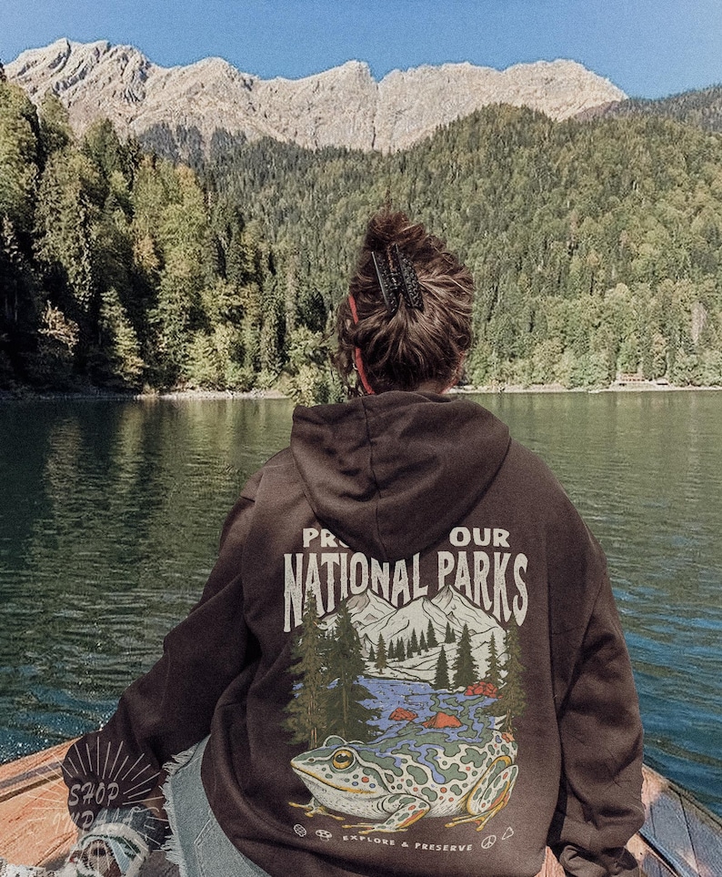 May include: A dark brown hooded sweatshirt featuring a graphic on the back with the text PROTECT OUR NATIONAL PARKS and EXPLORE & PRESERVE. The design includes a large frog, pine trees, and mountain peaks in shades of green, blue, and white.