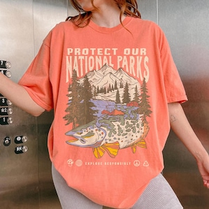 May include: A coral-colored t-shirt with a graphic print of a fish, mountains, and trees. The text "Protect Our National Parks" is printed on the shirt. The text "Explore Responsibly" is printed below the graphic.