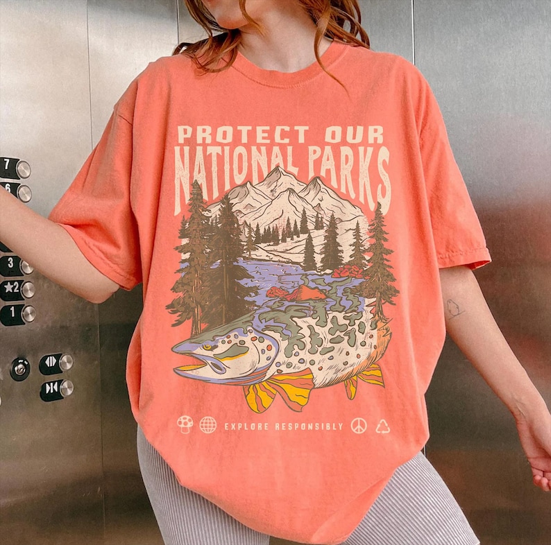 May include: A coral-colored t-shirt with a graphic print of a fish, mountains, and trees. The text "Protect Our National Parks" is printed on the shirt. The text "Explore Responsibly" is printed below the graphic.