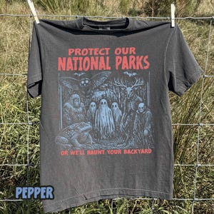 May include: A charcoal gray short-sleeve t-shirt featuring the red text PROTECT OUR NATIONAL PARKS and OR WE'LL HAUNT YOUR BACKYARD. The graphic depicts a moth, a toad, three ghosts, a deer-like creature, and a hooded figure in light blue ink.