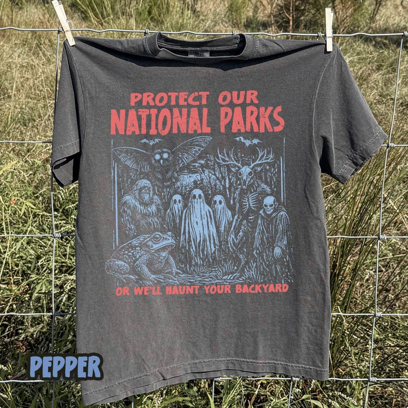 May include: A charcoal gray short-sleeve t-shirt featuring the red text PROTECT OUR NATIONAL PARKS and OR WE'LL HAUNT YOUR BACKYARD. The graphic depicts a moth, a toad, three ghosts, a deer-like creature, and a hooded figure in light blue ink.