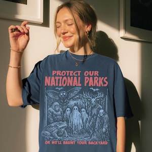 May include: A dark blue t-shirt featuring a graphic print with the text PROTECT OUR NATIONAL PARKS OR WE'LL HAUNT YOUR BACKYARD. The illustration depicts a toad, a large owl, a sasquatch, three ghosts, a deer-skulled creature, and a hooded figure.