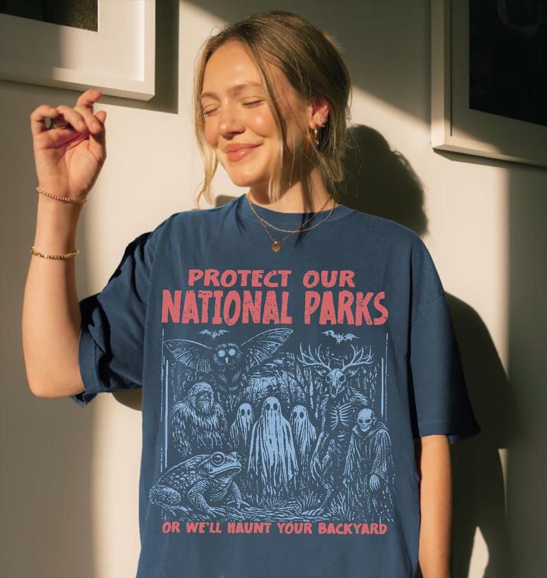 May include: A dark blue t-shirt featuring a graphic print with the text PROTECT OUR NATIONAL PARKS OR WE'LL HAUNT YOUR BACKYARD. The illustration depicts a toad, a large owl, a sasquatch, three ghosts, a deer-skulled creature, and a hooded figure.