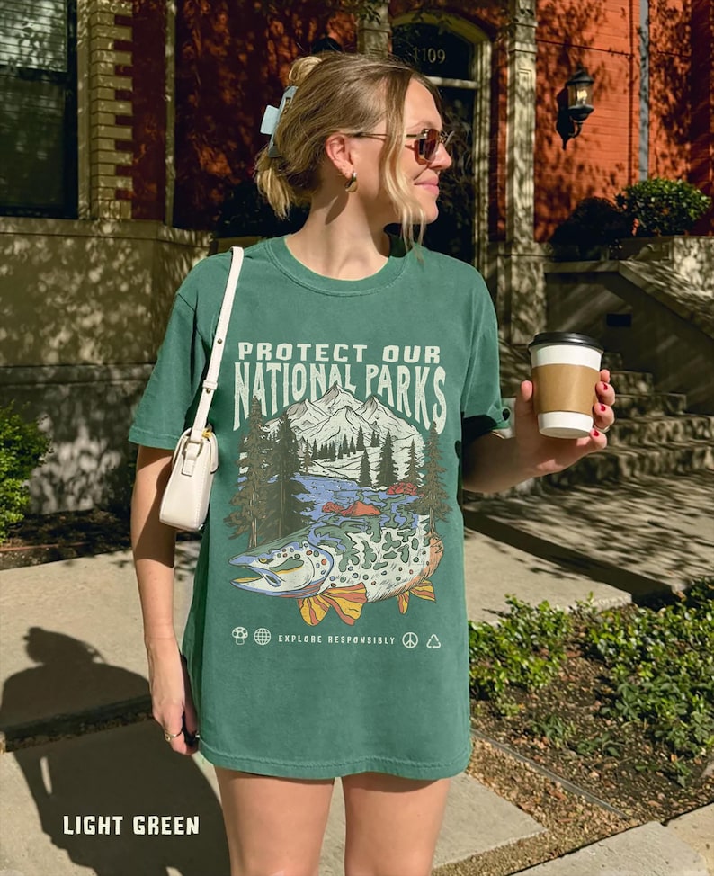 May include: A light green t-shirt with a graphic print of a mountain scene and a fish. The text "Protect Our National Parks" is printed on the shirt. The shirt also has the text "Explore Responsibly" printed on it.
